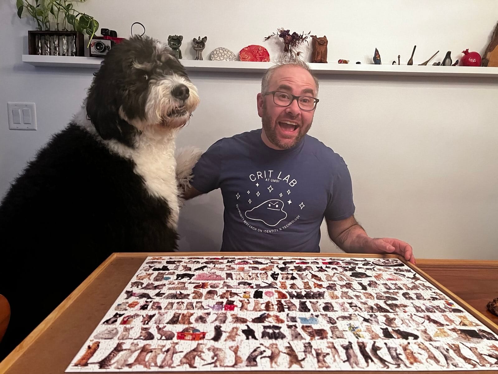 The author Oliver Haimson is seated next to his big dog at a table with a completed puzzle depicting dozens of cats.